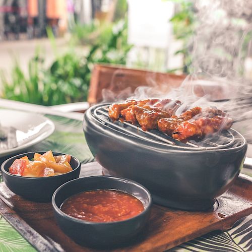 Grilled skewers on a hot plate, accompanied by two sauces and a side of rice on an outdoor table with a tropical-style tablecloth.