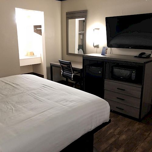 This image shows a hotel room with a bed, desk, chair, wall-mounted TV, drawers, and a vanity area with a mirror and lighting.