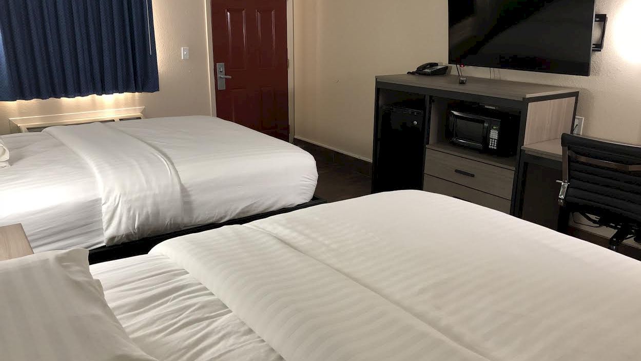 The image shows a hotel room with two neatly made double beds, a flat-screen TV, a desk, microwave, mini-fridge, and an open door.