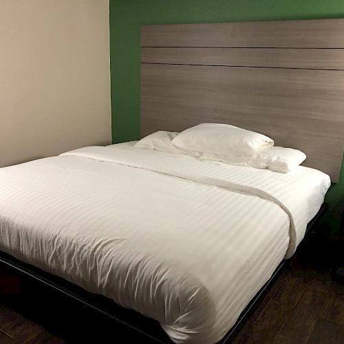 The image shows a neatly made bed with white bedding, a wooden headboard, and a nightstand on the right side, all in a room with green and beige walls.