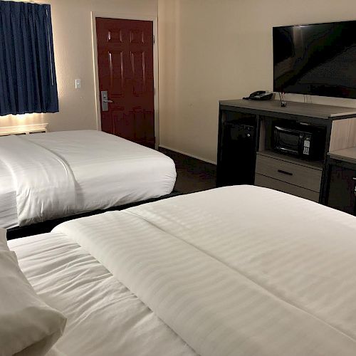 The image shows a hotel room with two double beds, a TV mounted on a dresser with a microwave and mini-fridge, a window with blue curtains, and a door.