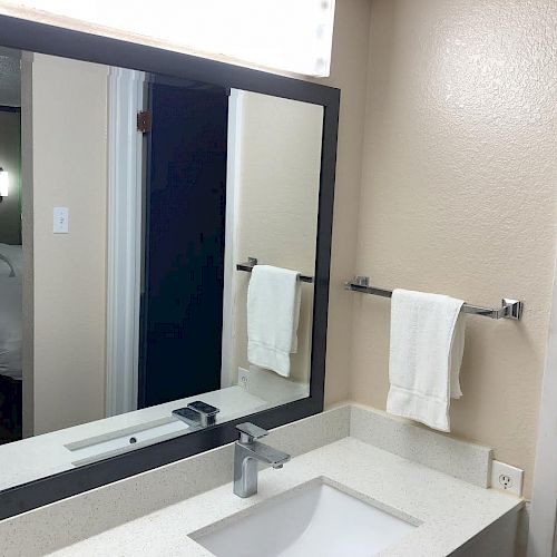 A modern bathroom with a large mirror, white sink, and faucet. A towel hangs on a rack to the right, reflecting a glimpse of a bedroom.