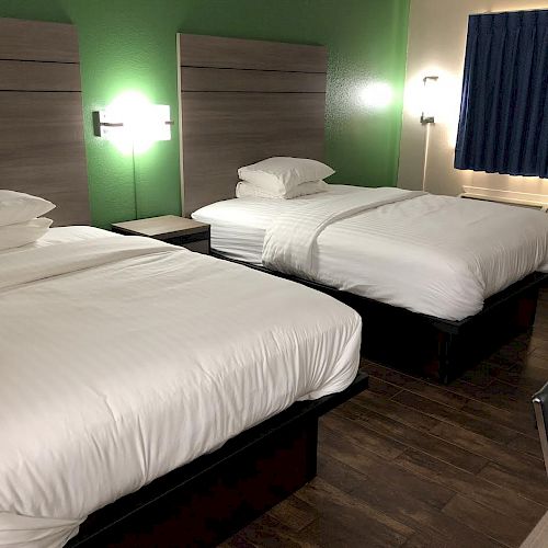 The image shows a hotel room with two beds, white bedding, a green accent wall, lamps, a window with blue curtains, and a chair at a desk.