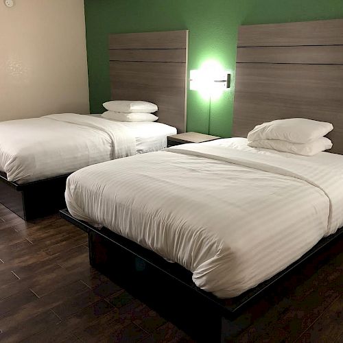 The image shows a neatly arranged hotel room with two beds, wood flooring, a green accent wall, and modern wall-mounted lights.