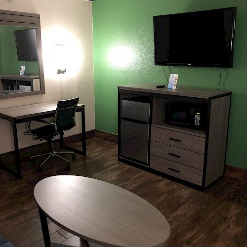 The image shows a hotel room with wooden flooring, a TV on the green wall, a desk with a chair, a mirror, and a cabinet with a microwave and mini fridge.