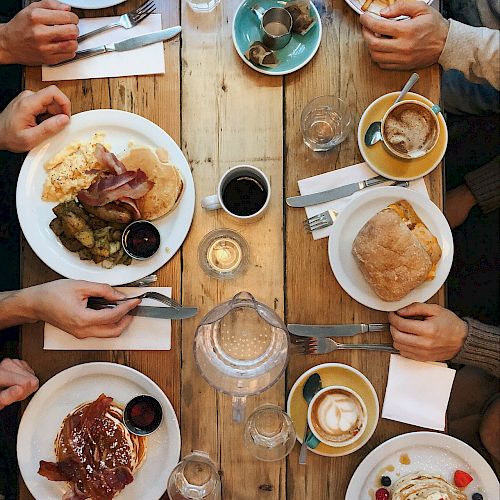 A top-down view of a wooden table with dishes, cups, glasses, and hands, suggesting a group meal or gathering.