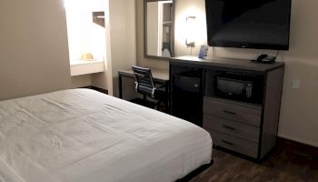 A modern hotel room with a neatly made bed, TV, desk, chair, and dresser in a clean, minimalistic design with a small bathroom area visible.