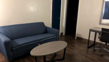 The image shows a room with a blue sofa, a wooden coffee table, a desk with a chair, and two doorways, all on a wooden floor.