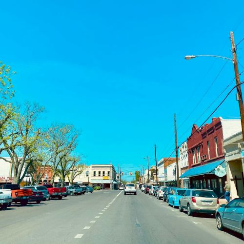 A small town street with cars parked on either side, colorful buildings, and trees against a clear blue sky.