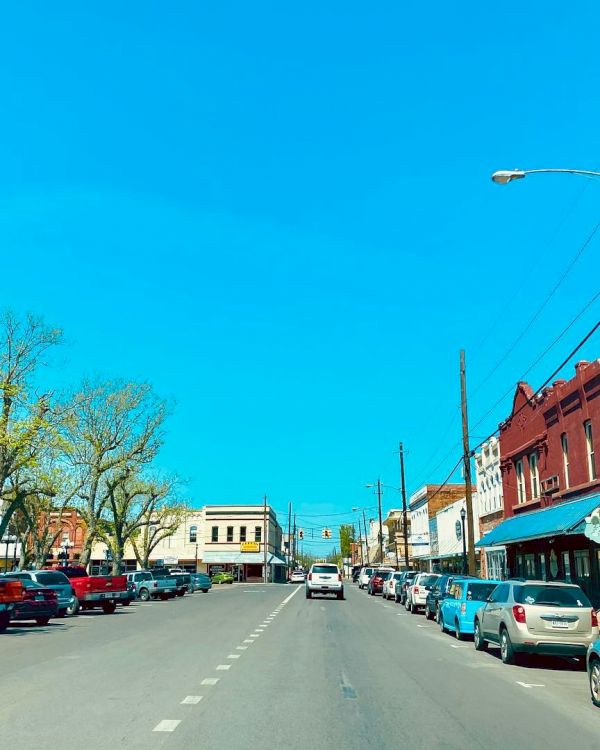 A small town street with cars parked on either side, colorful buildings, and trees against a clear blue sky.