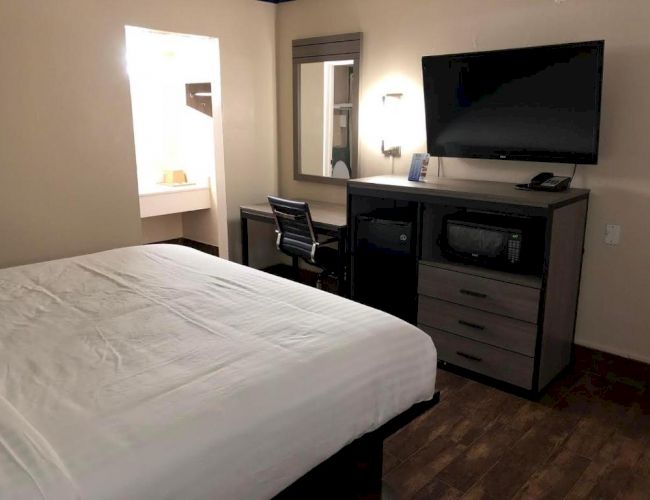 The image shows a hotel room with a bed, a desk with a chair, a wall-mounted TV, a mirror, and a small lamp.