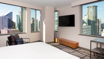 A modern hotel room with a bed, chair, TV, and large windows showcasing a cityscape with various high-rise buildings in the background.