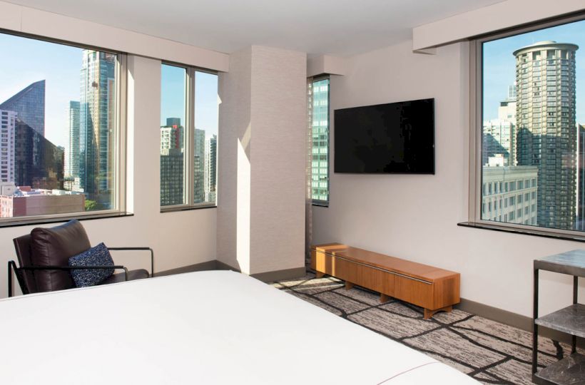 A modern hotel room with a bed, chair, TV, and large windows showcasing a cityscape with various high-rise buildings in the background.