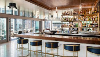 A modern bar with a well-stocked shelf, blue and gold stools, and floor-to-ceiling windows offering a view of the city, creating a sleek ambiance.