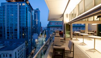 A modern cityscape from a balcony at dusk, featuring tall buildings, a street below, and an outdoor seating area with tables and chairs ending the sentence.
