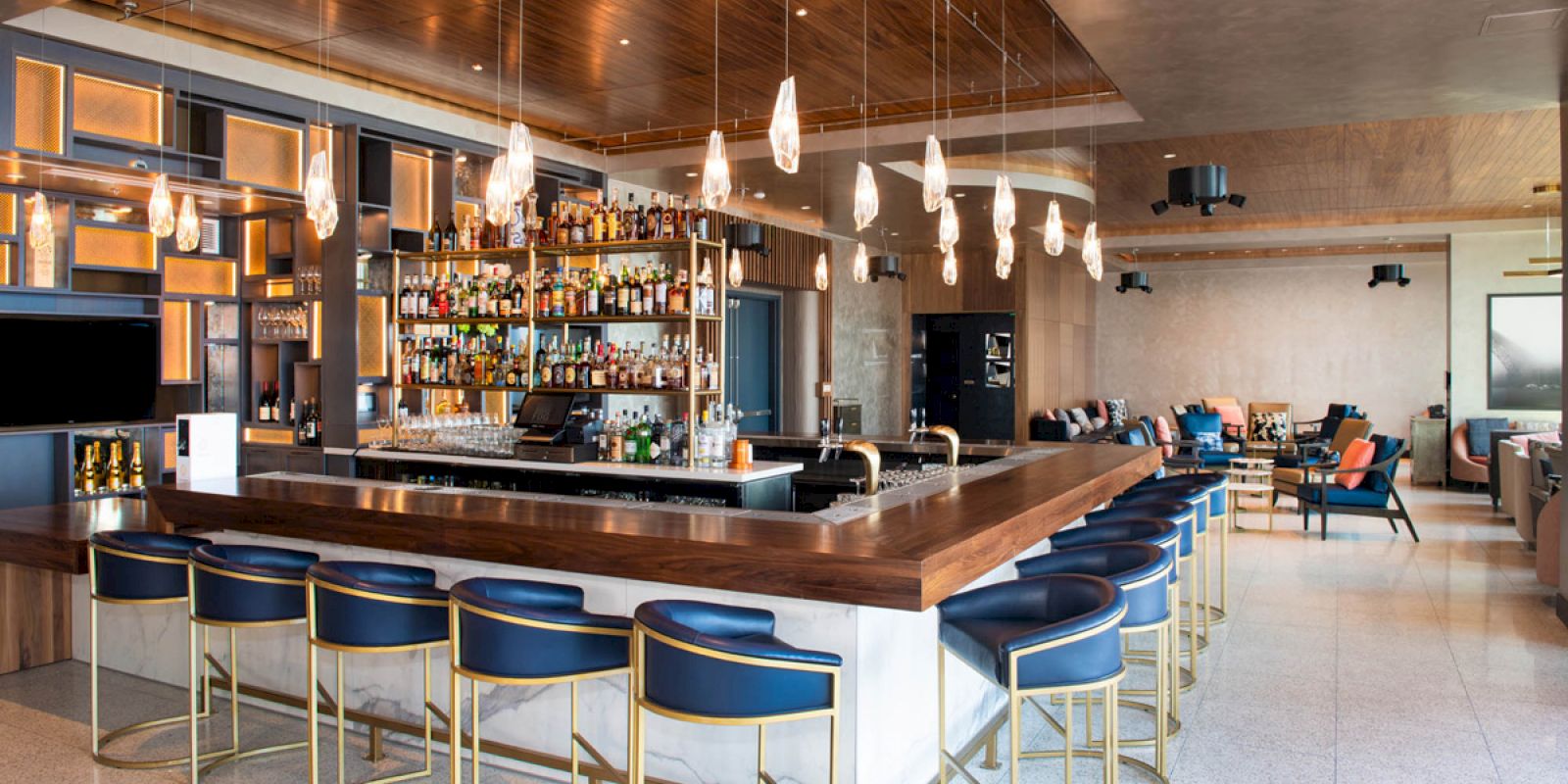 A modern bar with stylish blue and gold stools, a fully stocked bar, pendant lights, and a cozy seating area in the background.