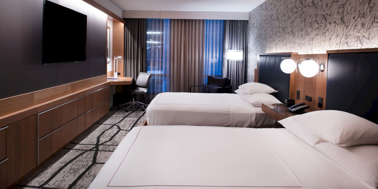 A modern hotel room with two neatly made beds, a wall-mounted TV, a work desk with a chair, and contemporary decor.
