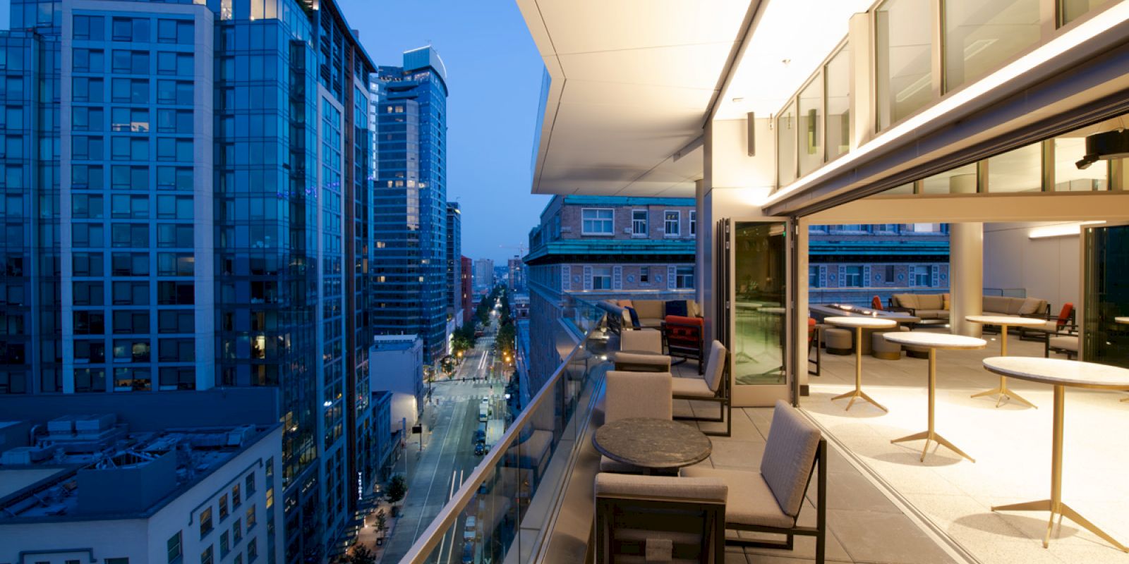An outdoor balcony at twilight, with tables and chairs, overlooking a city's glass skyscrapers and streets lit up in the evening glow.