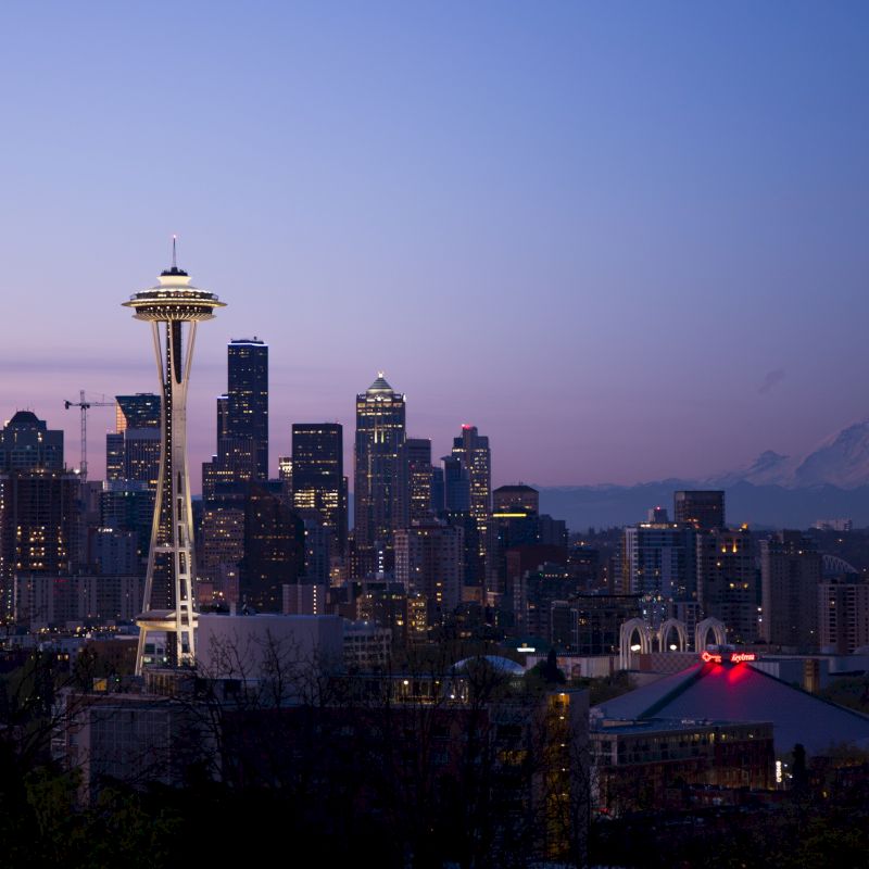 The image shows the Seattle skyline at dusk, featuring the iconic Space Needle, skyscrapers, and Mount Rainier in the background.