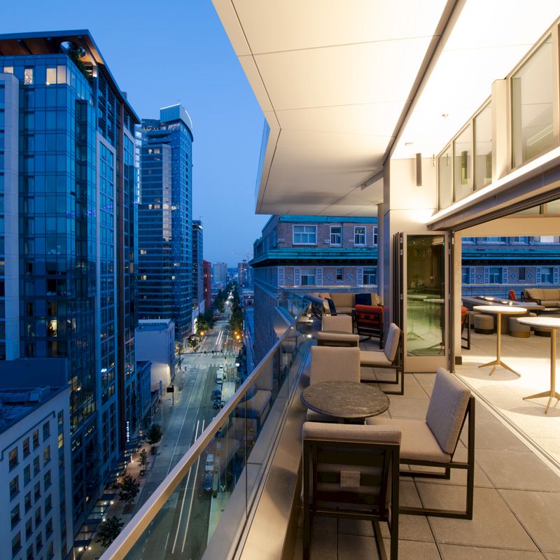 The image shows a modern cityscape from a high-rise balcony at dusk, with outdoor seating and tables, overlooking buildings and a street below.
