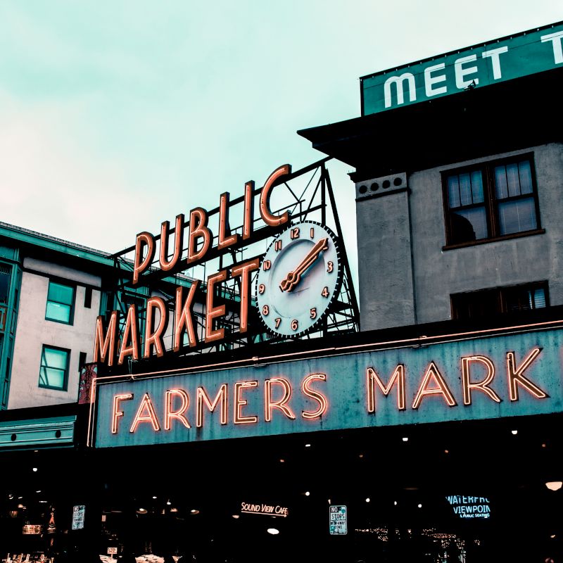 The image shows the iconic signs for a Public Market and Farmers Market, with a clock on the "MARKET" sign, at what appears to be a city market area.