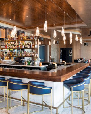 A modern, spacious bar with a wooden counter, blue chairs, and various hanging lights, featuring a well-stocked liquor shelf and a lounge area.