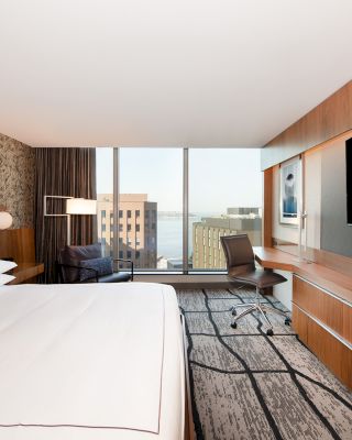 A modern hotel room with a large bed, wall-mounted TV, desk with chair, and a window offering a cityscape view.