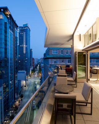 The image shows a modern urban balcony overlooking a city street with tall buildings. The balcony is furnished with tables and chairs.