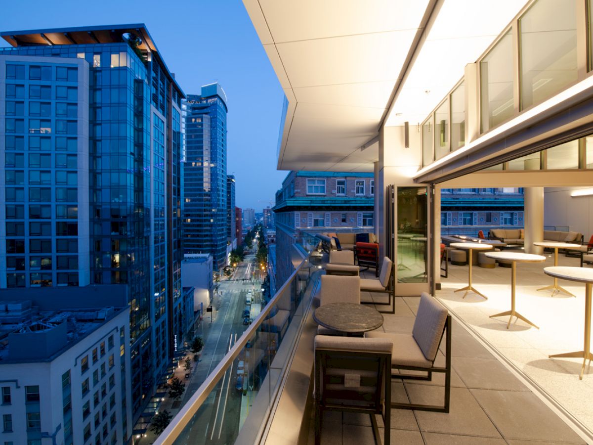 A modern rooftop terrace with seating and tables overlooking a city skyline at dusk, featuring high-rise buildings and streetlights below.
