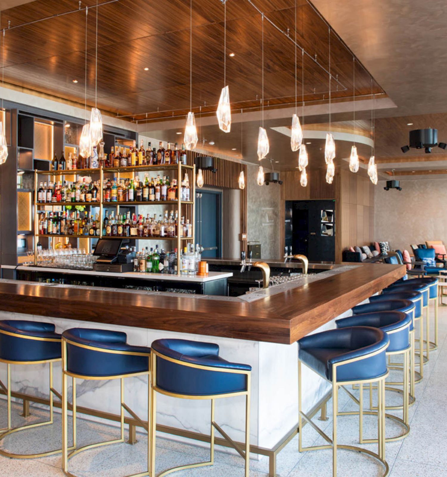 A modern, well-lit bar with blue and gold bar stools, a fully stocked liquor shelf, hanging lights, and a seating area in the background.