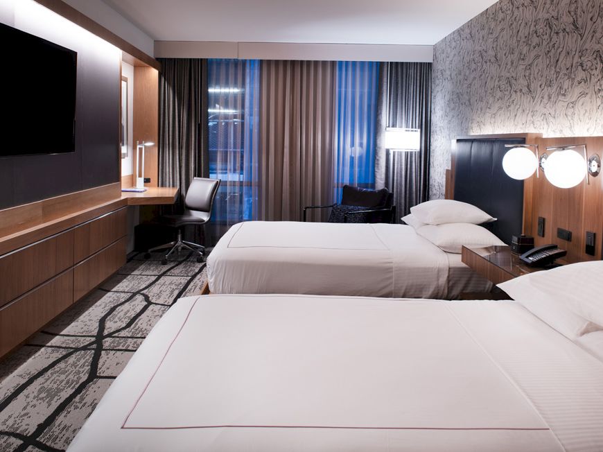 The image shows a modern hotel room with two beds, a TV, a work desk, and a large window with curtains. The decor is contemporary and clean.