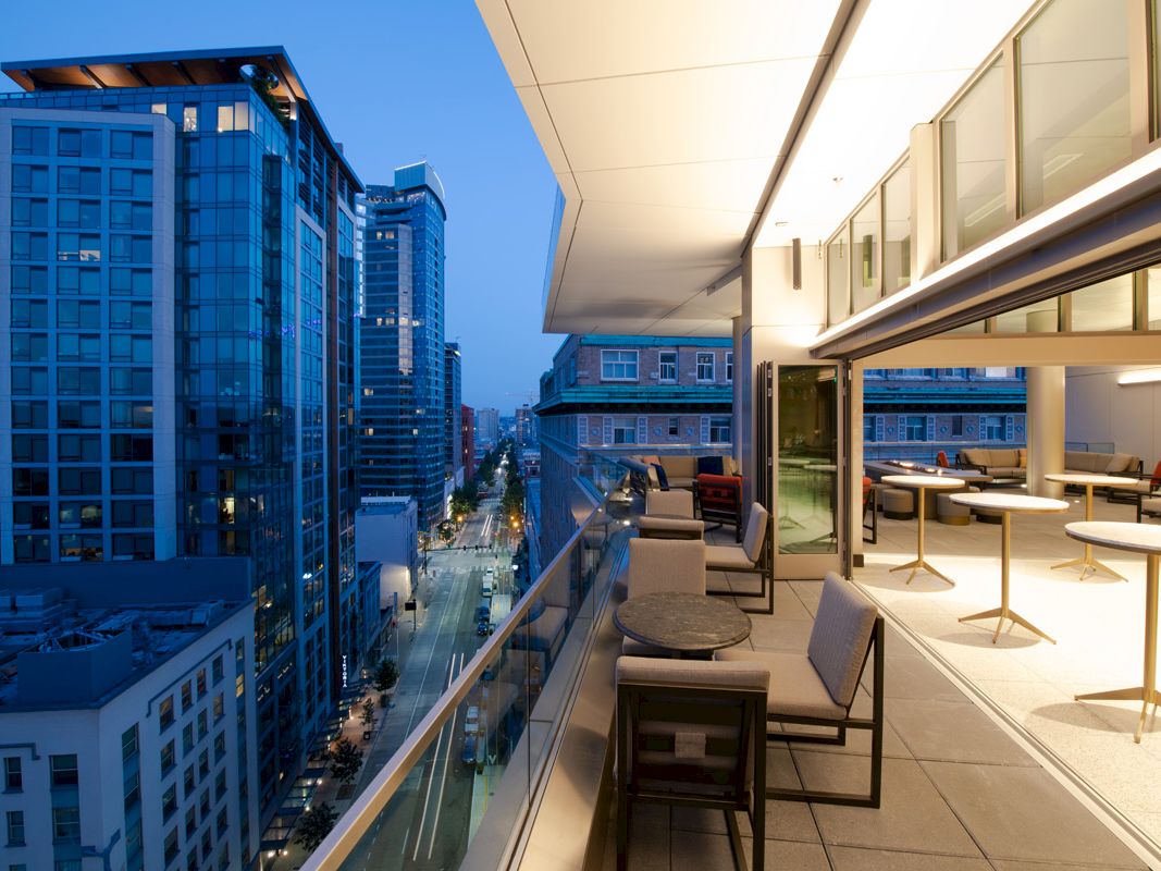 The image features a modern balcony setting with tables and chairs, overlooking a cityscape filled with tall buildings at dusk.