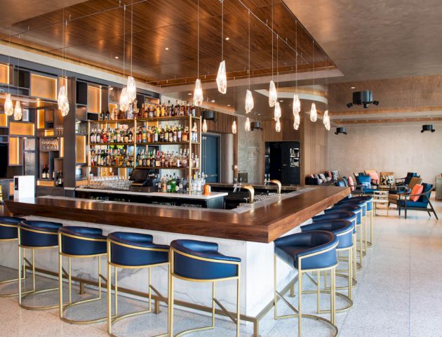 A modern bar with a wooden counter, blue bar stools, pendant lights, stocked shelves, and a seating area in the background.