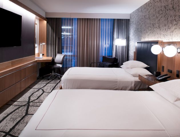 A modern hotel room features two neatly made beds, a wall-mounted TV, a desk with a chair, and stylish decor with patterned wallpaper and carpet.