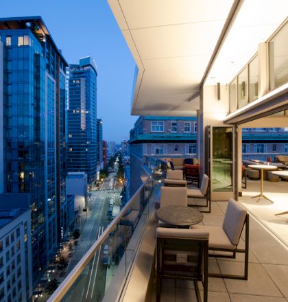 The image shows a high-rise balcony with chairs and tables overlooking a city street lined with modern buildings and lit up at dusk.
