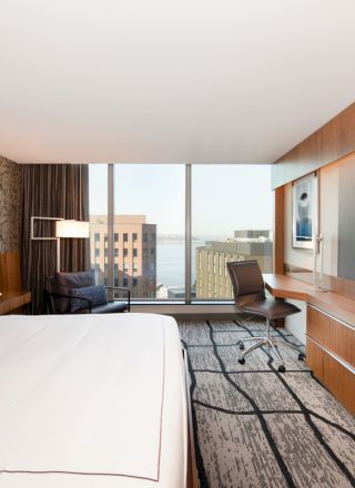 This image shows a modern hotel room featuring a large bed, a wall-mounted TV, a desk, and a window with a cityscape view in the background.