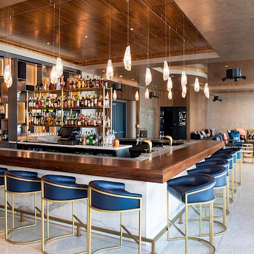 A modern bar with a large selection of bottles, blue bar stools, hanging lights, and a comfortable seating area in the background.