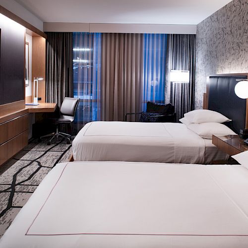 A modern hotel room with two neatly made beds, a wall-mounted TV, a desk and chair by the window, and stylish lighting and decor.