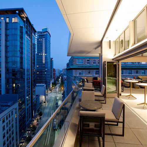 A modern balcony with seating overlooks a cityscape of high-rise buildings at dusk, featuring illuminated office spaces and a busy street below.
