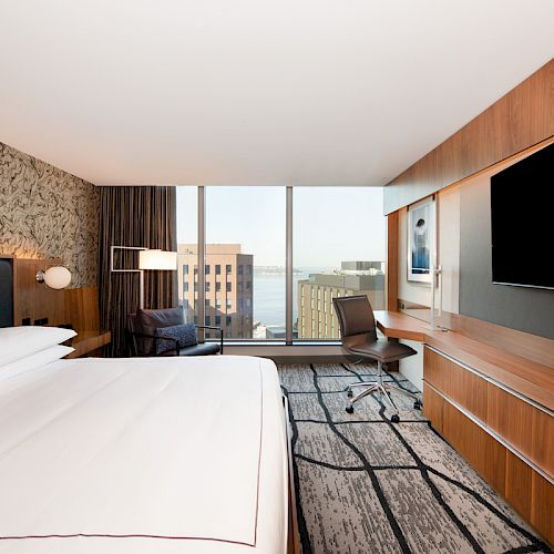 A modern hotel room with a large bed, desk, chair, flat-screen TV, and a window offering a cityscape and water view.