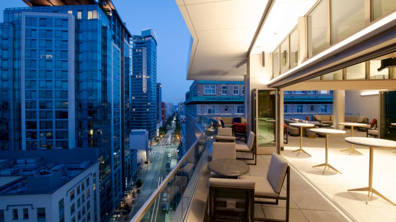 The image shows a modern cityscape viewed from a balcony with outdoor seating and tables. Tall buildings are in the background.
