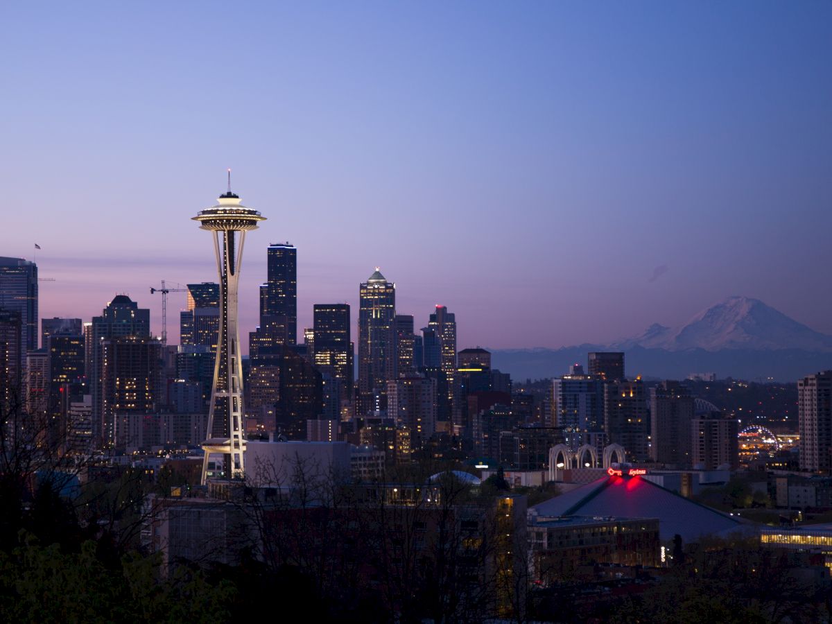 The image shows the skyline of Seattle at dusk, with the Space Needle prominently visible and Mount Rainier in the background.