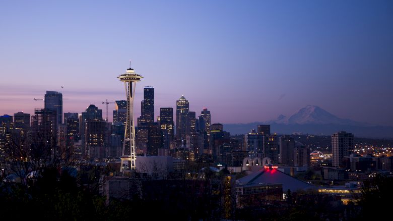 The image shows the skyline of Seattle at dusk, with the Space Needle prominently visible and Mount Rainier in the background.