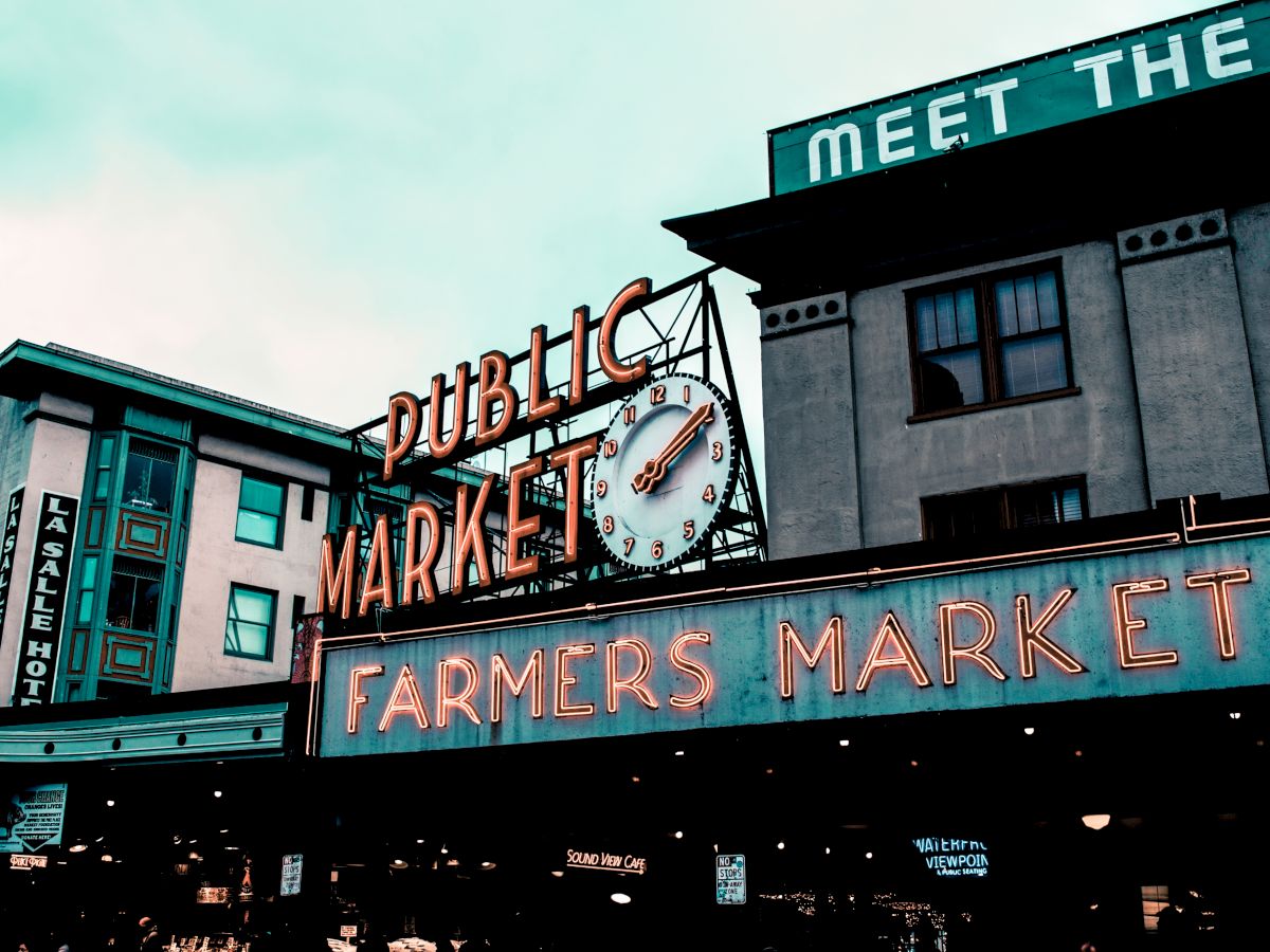 The image shows a neon sign for a "Public Market" and "Farmers Market" on a building. A clock is also part of the signage. The venue appears busy.