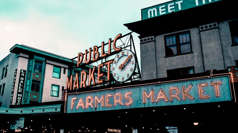 The image shows a neon sign for a "Public Market" and "Farmers Market" on a building. A clock is also part of the signage. The venue appears busy.