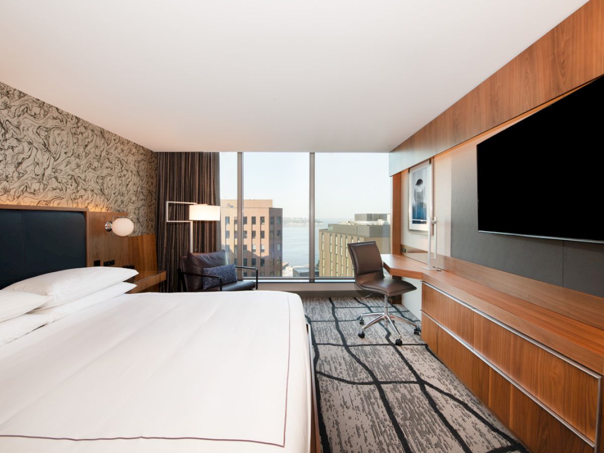 A modern hotel room with a large bed, desk, chair, TV, and a window with a city view is shown. The decor includes wood paneling and patterned wallpaper.