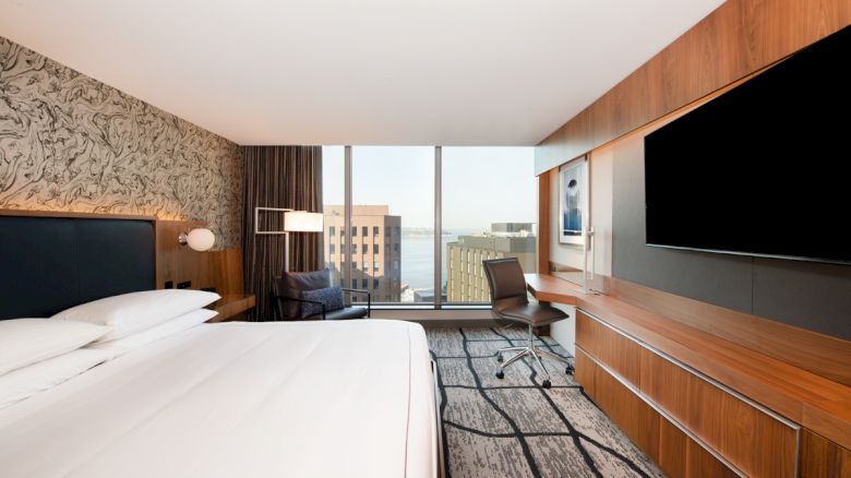 A modern hotel room with a large bed, desk, chair, TV, and a window with a city view is shown. The decor includes wood paneling and patterned wallpaper.