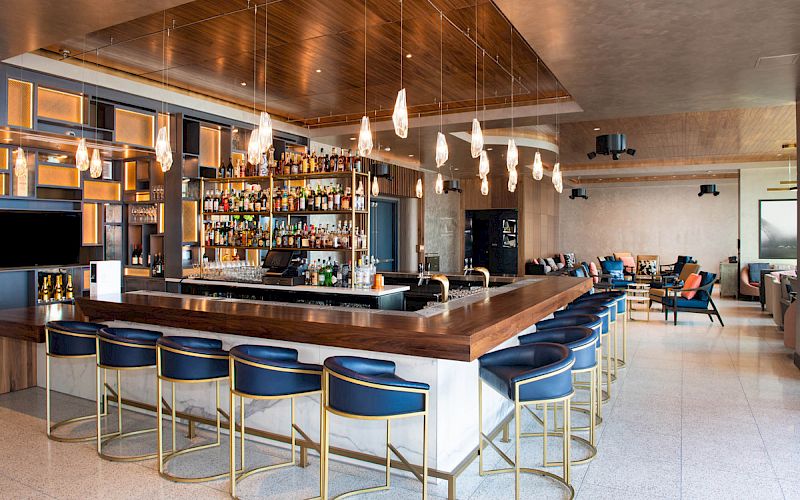 The image shows a modern bar with a counter surrounded by blue stools, a ceiling with pendant lights, and a well-stocked shelf of bottles.