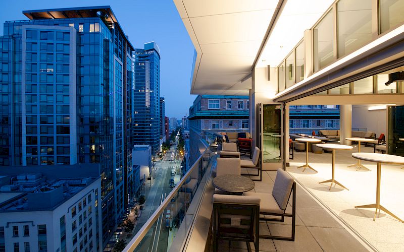 A modern balcony with seating areas overlooks a cityscape featuring tall buildings, city lights, and a clear evening sky.
