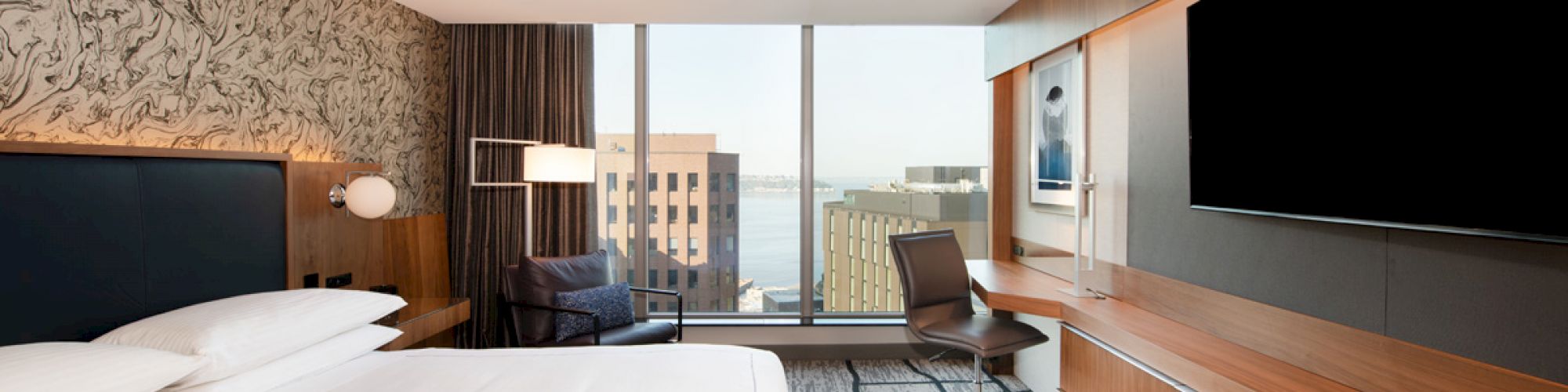 A modern hotel room features a large bed, wall-mounted TV, desk, chair, and a large window with a city and water view.